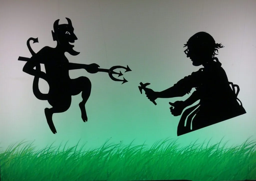 Personnages, ombres noires avec fils, qui descendent par dessus un écran lumineux, vert fluo : ici un diable avec sa fourche et un cordonnier avec son marteau.
