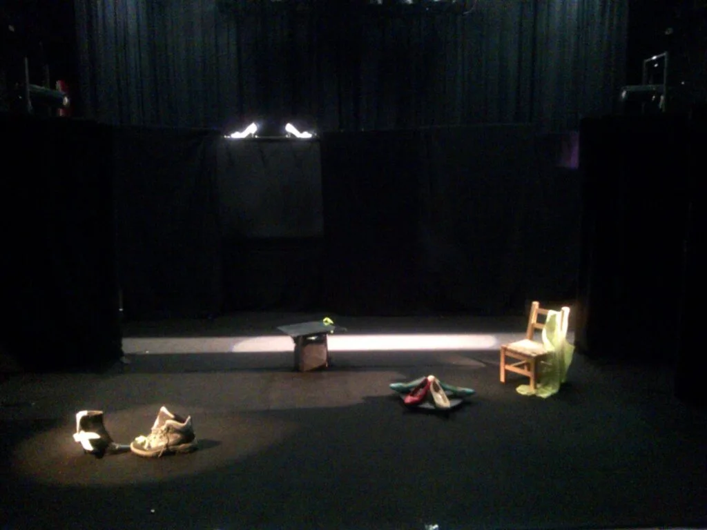 Le plateau dans un théâtre avant l’arrivée du public. Le tour de scène est réalisé avec des tentures noires. Au milieu, au lointain, on voit deux escarpins blancs qu’on devine qu’ils sont posés sur une tenture en hauteur. Une bande de lumière blanche traverse toute la scène au lointain. Devant cette bande vers le bord de scène, au sol, sont posés une chaise avec un vêtement vert sur le dossier, un miroir, un tourniquet de chaussures, puis plus près encore une grande basket et une bottine dans un rond d’éclairage.
