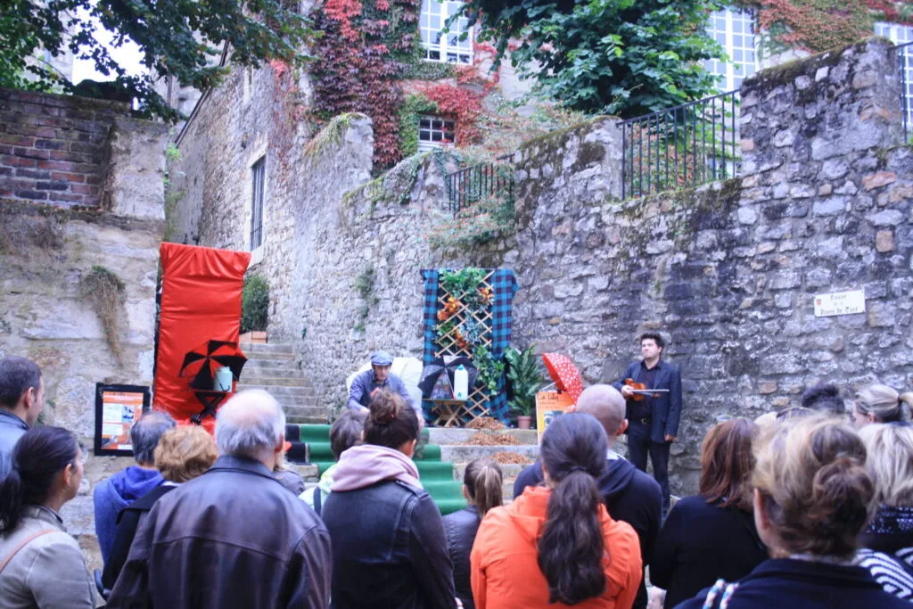 Décor posé sur le haut des marches en pierres grises avec un panneau rouge, un bidon de lait, un panneau à carreaux verts décoré de fleurs, un bidon d'eau. En bas de ces escaliers, un public d'adultes. La marionnette est aussi pour les adultes