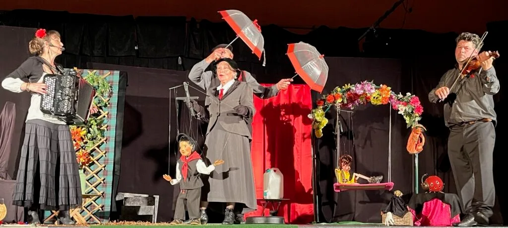 Spectacle en salle. Tableau avec la chanson "Chem chem Cheminée" accompagnée d'une accordéoniste, d'un violoniste. Au centre, le comédien avec ses parapluies ouverts sur la marionnettiste, la nouvelle marionnette, le petit Ramoneur.