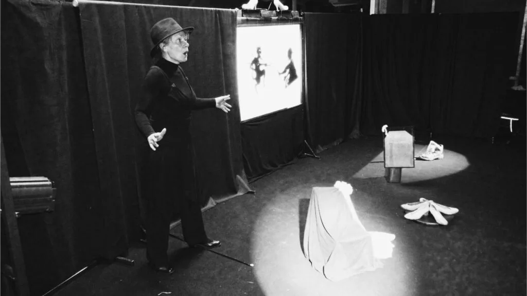 Image en noir et blanc avec Liliane Plouzeau, la bouche formulant un Oh ! de surprise, répète devant un public fictif. Au milieu de tentures noires, un écran avec un diable et un cordonnier en théâtre d’ombres. Devant divers accessoires