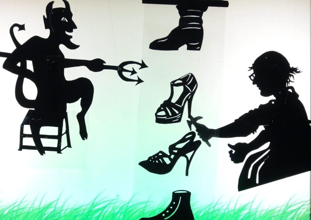 Personnages en ombres posés sur un écran lumineux vert. Le diable pointe sa fourche vers le cordonnier qui répare avec son marteau une des chaussures qui descendent du haut vers le bas devant lui.