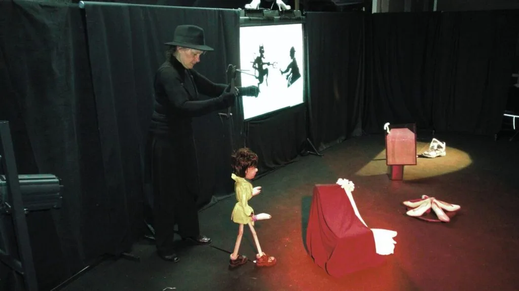 Valentine la marionnette à fils haute de 80 cm, de jaune vêtue arrive sur scène manipulée par la marionnettiste tout en noir, chapeau compris. Le décor est constitué de tentures noires, d'un théâtre d'ombres, d'un fauteuil, des chaussures.