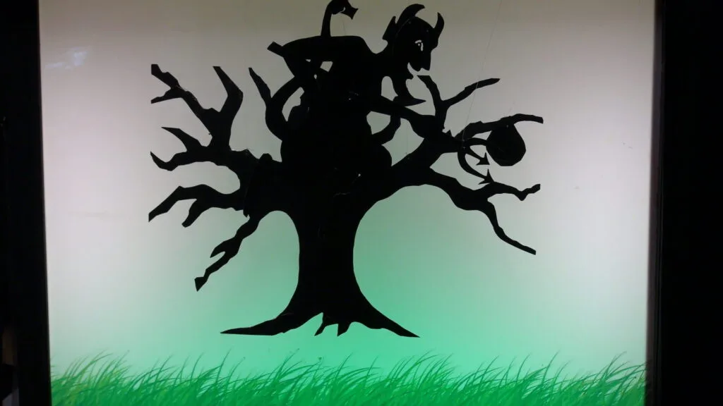 Théâtre d’ombres: un diable avec sa fourche, sur la cime d'un arbre, essaie d'attraper une poire. Le diable et l'arbre, silhouettes en noir sont tenues par des fils qui descendent du dessus pour être posés sur l'écran lumineux vert fluo.