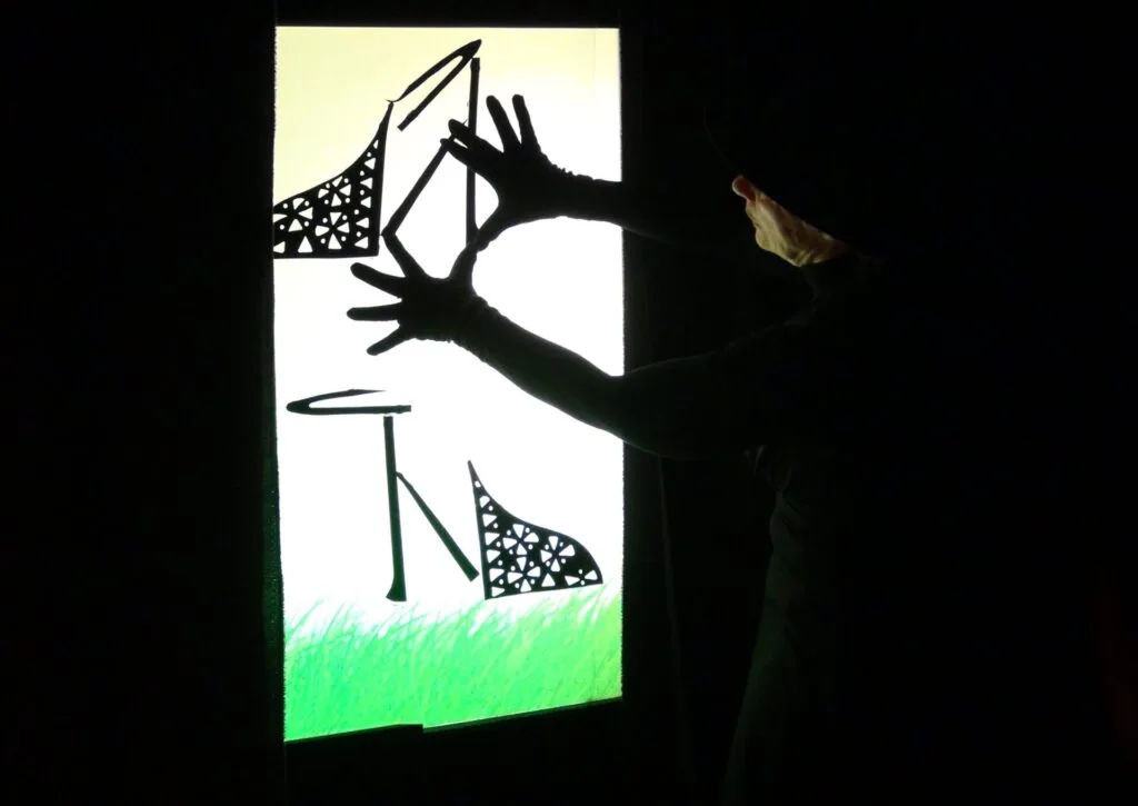 Suite à l'image ci-avant, la marionnettiste a transformé la coupe en cristal en deux escarpins avec les morceaux noirs qu'elle colle ou décolle sur l'écran lumineux vert fluo. On aperçoit les mains et discrètement le visage de la marionnette.