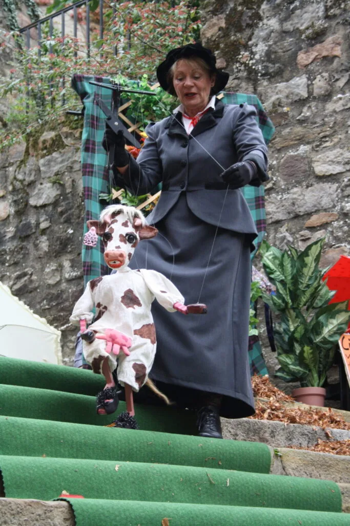 Gigi la vache, la marionnette à fils danse avec la marionnettiste sur une marche d'un escalier en pierres sur lequel est déroulé un tapis vert. Un grand mur en pierres bordé de feuillages longe cet escalier.