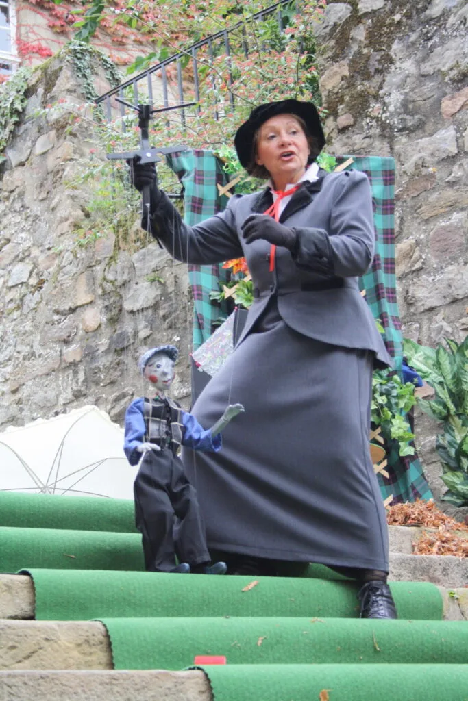 La marionnettiste avec un costume gris style Mary Poppins chante avec sa marionnette le petit ramoneur, vêtu de gris aussi. Il regarde devant lui, debout. Tous les deux sont dans un escalier en pierres et tentent de descendre une marche.