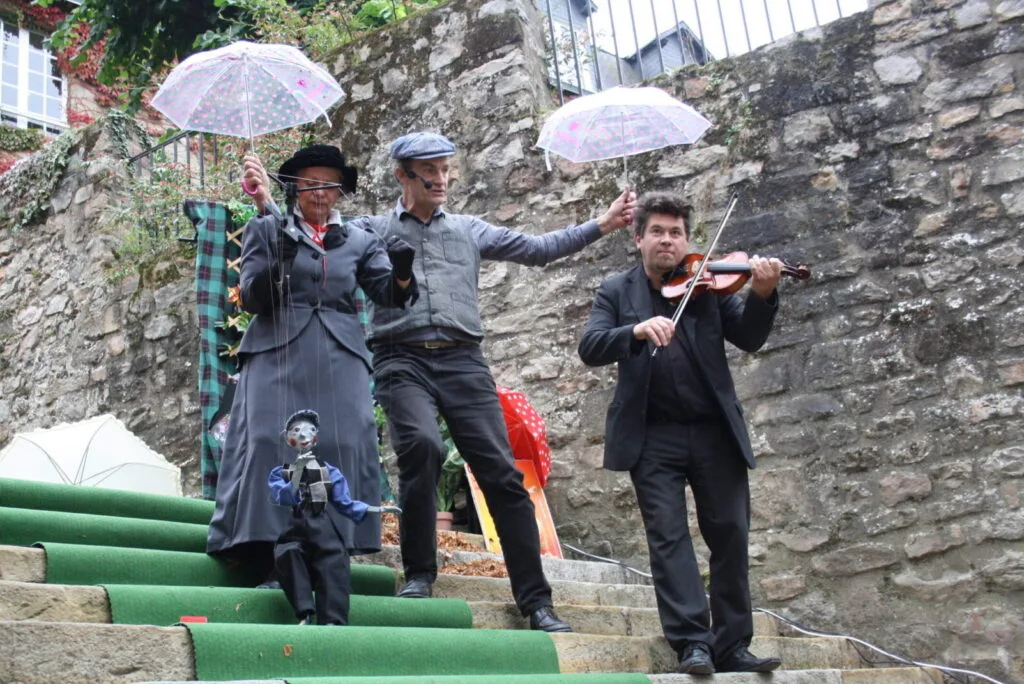 La marionnettiste manipule la marionnette, le petit ramoneur, le violoniste joue, le comédien déploie deux petits parapluies au dessus d'eux. Tous descendent l'escalier de pierres sur un tapis vert. Tous sont vêtus de gris.
