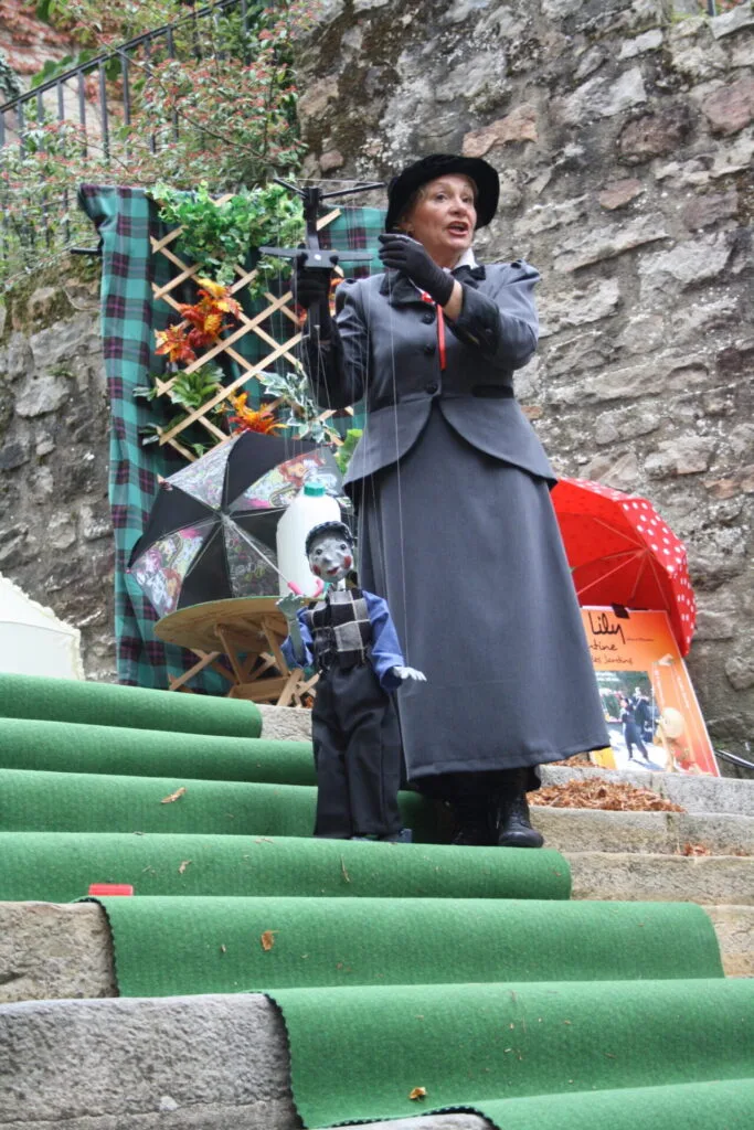 La marionnettiste chante avec sa marionnette le petit ramoneur. Ils sont sur un tapis vert déroulé sur les marches d'un escalier en pierres avec derrière eux une partie du décor. Un grand mur en pierres bordé de feuillages longe cet escalier