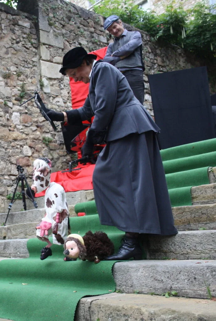 La marionnettiste et les deux marionnettes à fils qu'elle manipule, seule, descendent les marches d'un escalier en pierres recouvert d'un tapis vert. Derrière elles, le comédien regarde, en souriant, Gigi la vache et le hérisson.