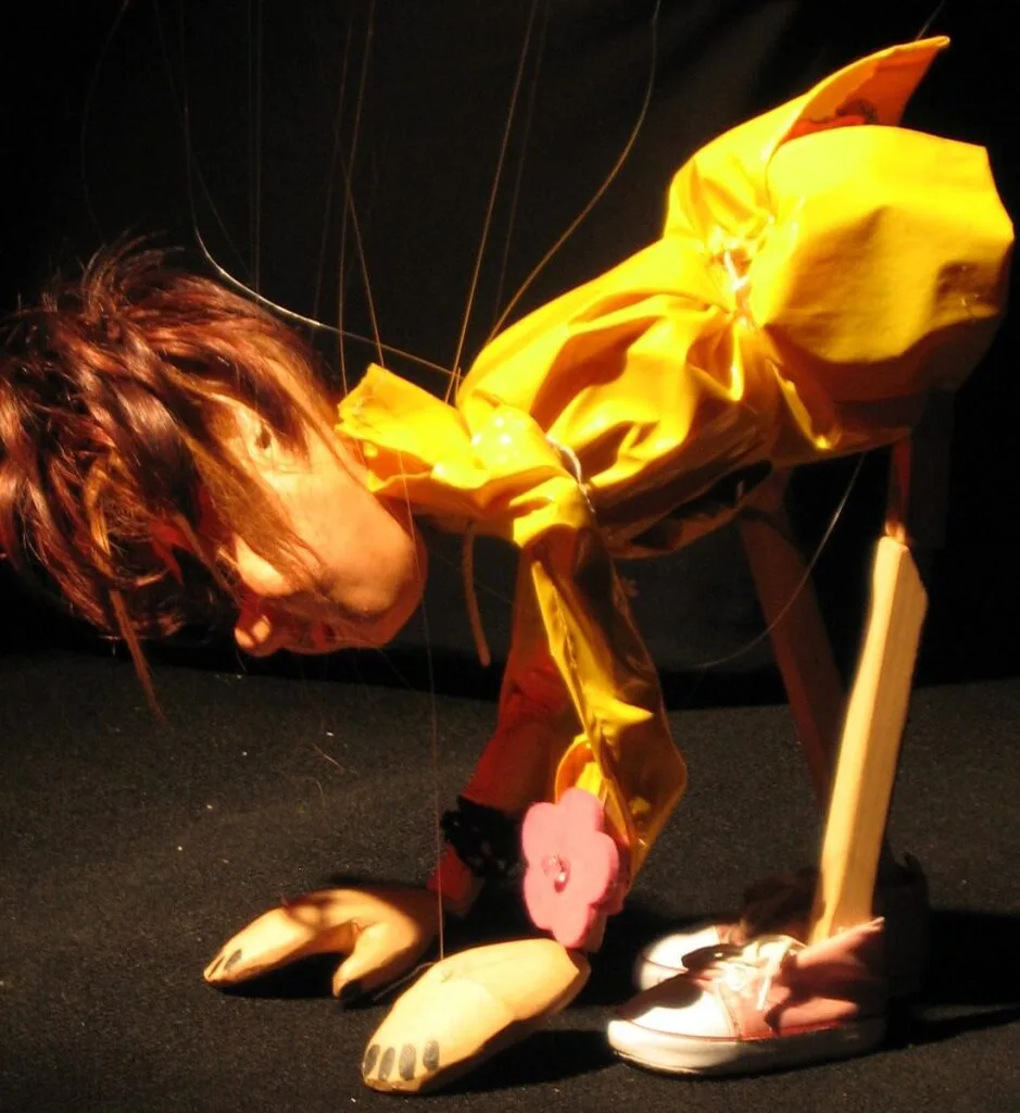 Valentine, la marionnette fétiche de la Compagnie Lily, se délasse avec quelques étirements : le dos arrondi, elle touche le sol de ses mains, les jambes tendues. Elle est habillée d’un ciré jaune, et porte des baskets roses.