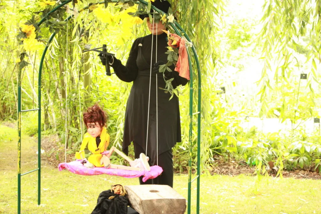 Valentine la marionnette à fils, vêtue de jaune est assise sur sa balançoire. Derrière elle, la marionnettiste en noir. Au premier plan, on voit le bourreau près de son billot. Sur fond de verdure, le tableau est d'un vert tendre lumineux.