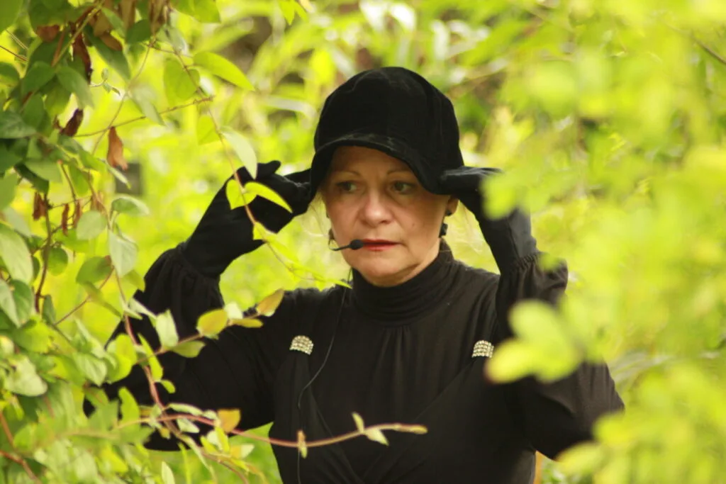 On voit le buste d'une femme, Liliane Plouzeau, vêtue de noir, Ses mains gantées de noir sont posées sur son chapeau noir. Elle fixe attentivement un point. Elle a un micro HF devant sa bouche mais elle ne dit rien. Ce qui est beau dans cette image, c'est le buste noir de cette femme sur un fond de feuillage vert très clair, très printenanier.