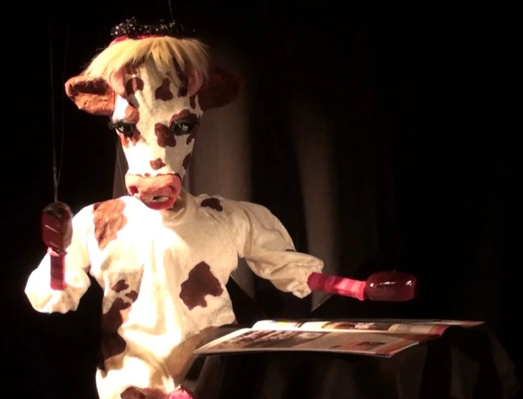 La marionnette Gigi la vache, un sabot marron brillant sur une revue déposée sur la table, l’autre sabot levé en l'air, porte une robe blanche aux tâches marron avec des manchons roses vif, des petites cornes avec un noeud, son museau est rose