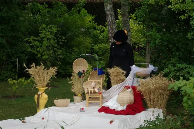 Décor avec des tiges de blé, des corbeilles en osier, une marionnette assise sur une chaise, une autre allongée sur un tissu rouge, le tout sur un long tissu blanc sur fond d'arbres vert foncé. On aperçoit discrètement la marionnettiste.