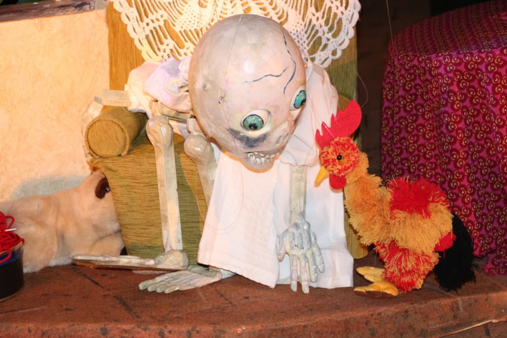 Gros plan sur la Mort malade qui regarde, penchée tout de travers, la marionnette, la poule rouge orangé, de Madeleine