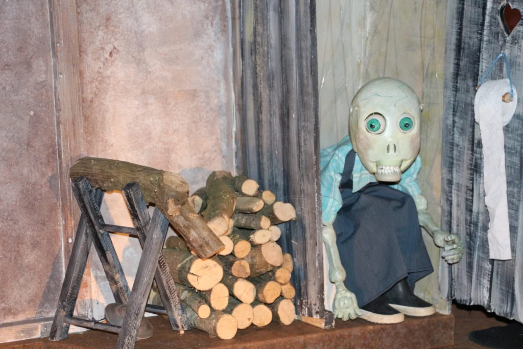 La marionnette représentant la Mort habillée en fermière avec ses bottes est assise sur des WC car un rouleur de papier pend. A côté, il y a un tas de bois. La Mort parait surprise avec ses gros yeux verts.