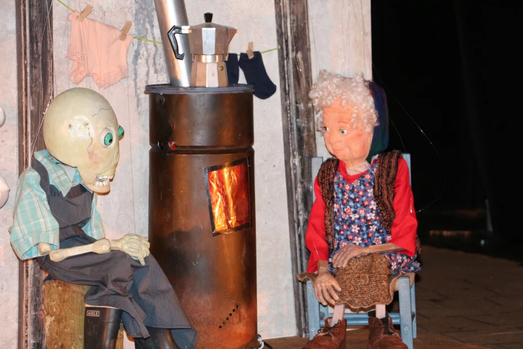 Gros plan sur la scène de la veillée au coin du feu avec les marionnettes à fils, Madeleine et la Mort habillée en fermière. Papotage . Le café chauffe sur le poêle.