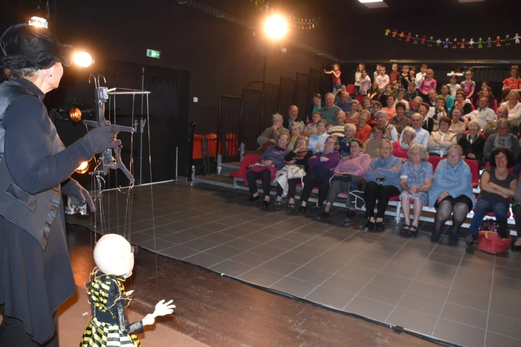 Liliane Plouzeau présente la marionnette Mme La Mort avec sa robe à rayures jaunes et noires à un public assis dans des gradins, dans un théâtre, Un public de personnes âgées et une classe d'enfants d'école primaire.