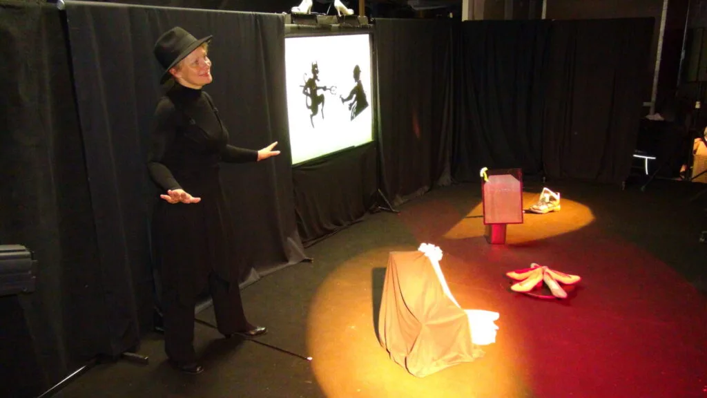 Liliane Plouzeau, vêtue de noir, s’adresse en souriant à un public imaginaire dans son atelier où devant des tentures noires se trouvent un théâtre d'ombres, des accessoires, fauteuil, chaussures, dans un éclairage jaune orangé. Tableau 1 sur les 4 suivants