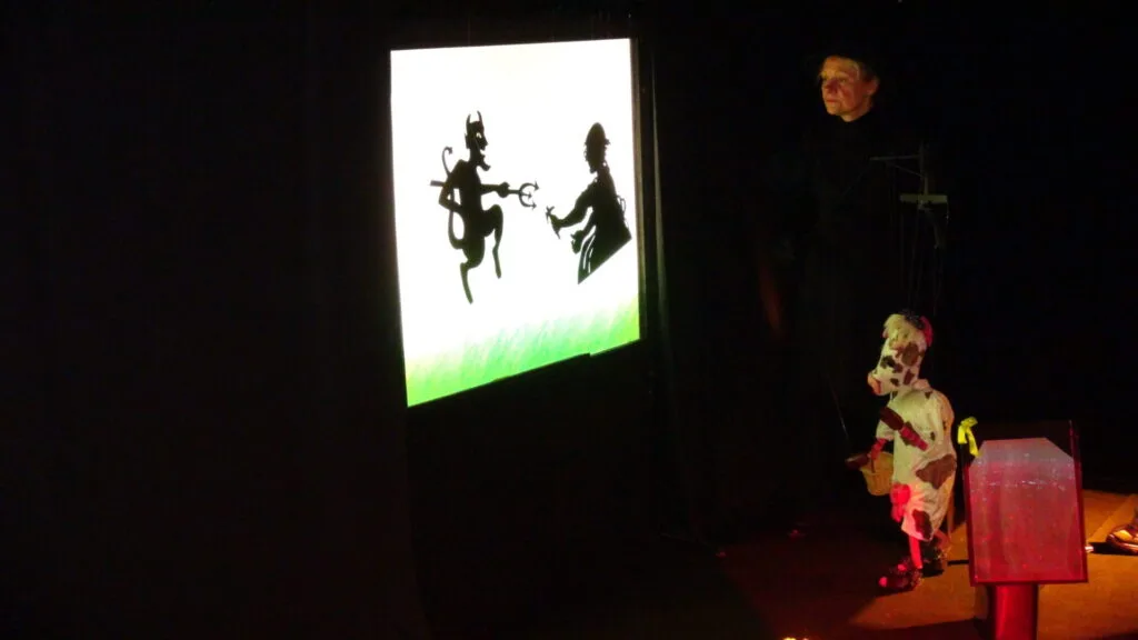 La marionnette Gigi la vache est debout devant un écran. Elle regarde le diable avec sa fourche et le cordonnier, tous deux représentés en théâtre d’ombres. On distingue dans la pénombre le visage de la marionnettiste.