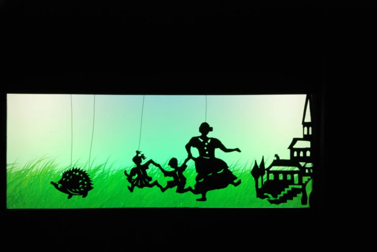 Théâtre d’ombres : sur un écran vert fluo, plusieurs silhouettes découpées apparaissent suspendues. Un hérisson stylisé, puis trois enfants se tenant la main avec leur maman courent en se suivant. Au loin, un village avec un clocher