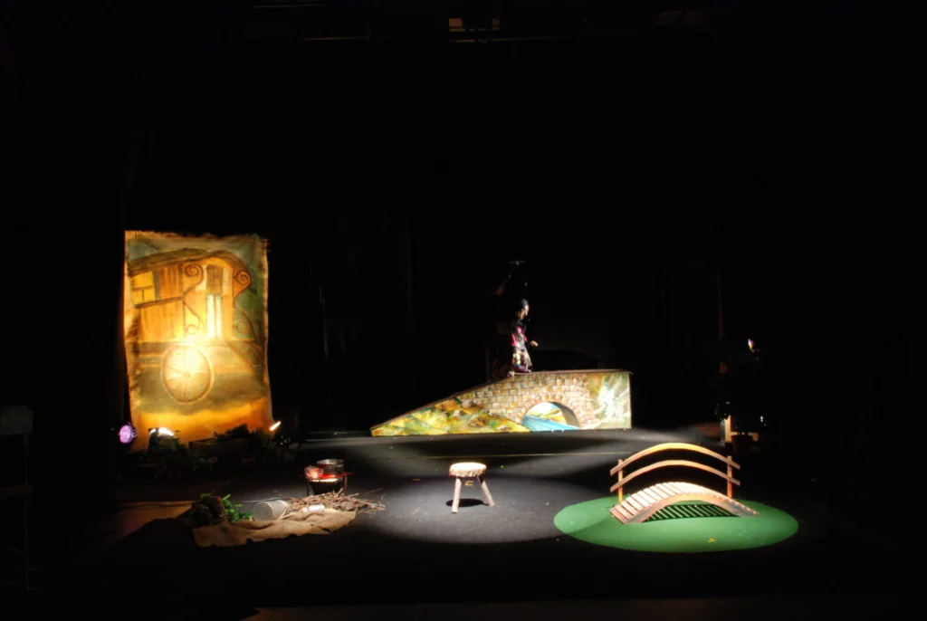 Décor épuré et contemporain, roulotte peinte, chemin, feu de camp et ponts mis en valeur par des points lumineux sur fond de scène noir. Une marionnette monte le chemin au lointain