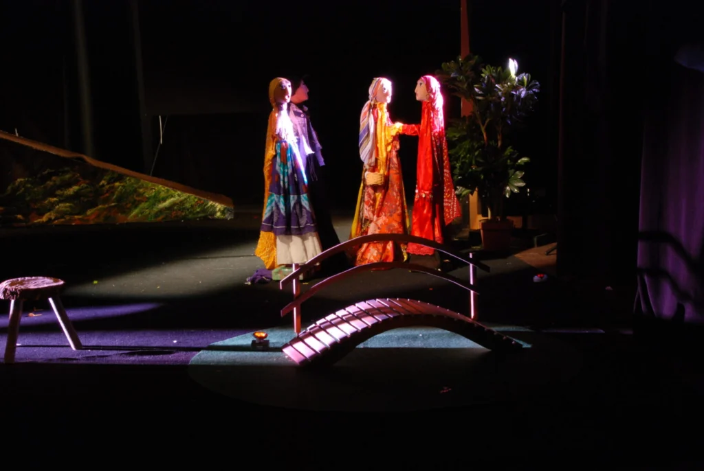 Sur fond de scène noir, trois marionnettes sur pied, en robes colorées sont près d’un petit pont. Une lumière chaude éclaire leurs visages. Au lointain on voit un début de chemin qui monte. Des faisceaux de lumière douce évoquent la nuit.
