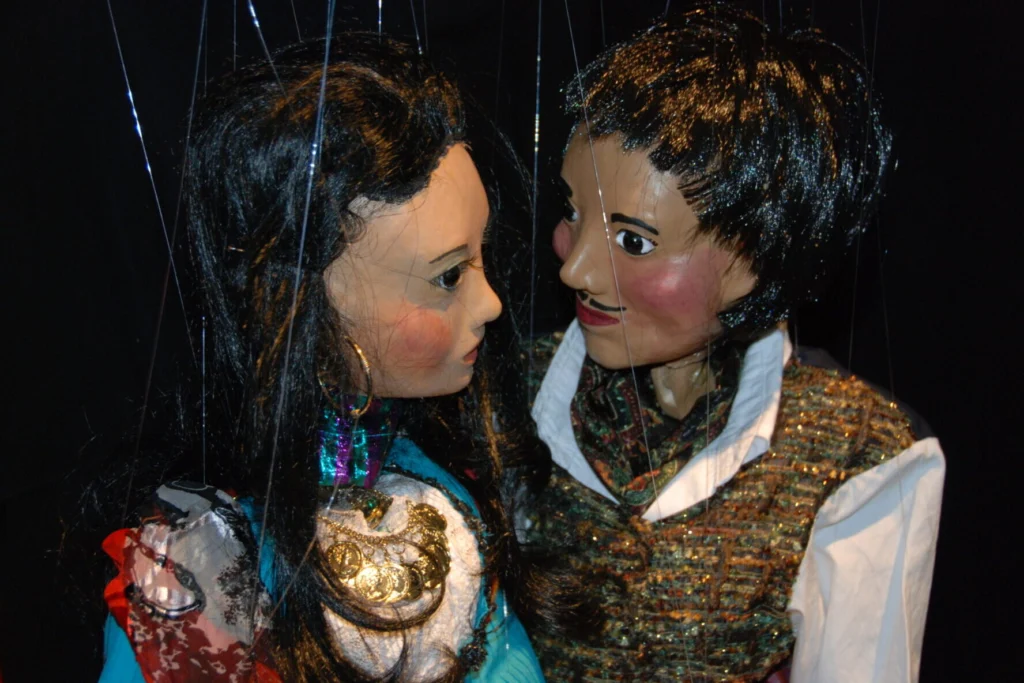 Portait serré du buste deux marionnettes à fils tziganes. Un jeune garçon vêtu d'un gilet scintillant regarde intensément une jeune fille au chemisier coloré avec des bijoux. Leurs deux visages rapprochés ne sont que tendresse.