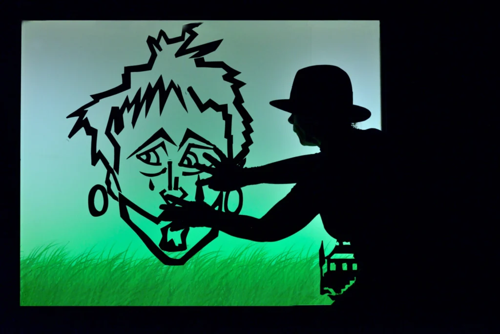 « Théâtre d’ombres sur un écran lumineux vert, où la marionnettiste en noir façonne la silhouette d’un visage tzigane projeté en ombre.