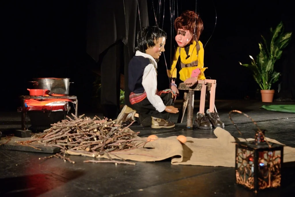 Deux marionnettes à fils discutent près d’un petit feu de camp. Le garçon tzigane agenouillé s’adresse à la jeune fille vêtue de jaune assise sur un tabouret. Autour d’eux, bois, casseroles et lampe jonchent le sol.