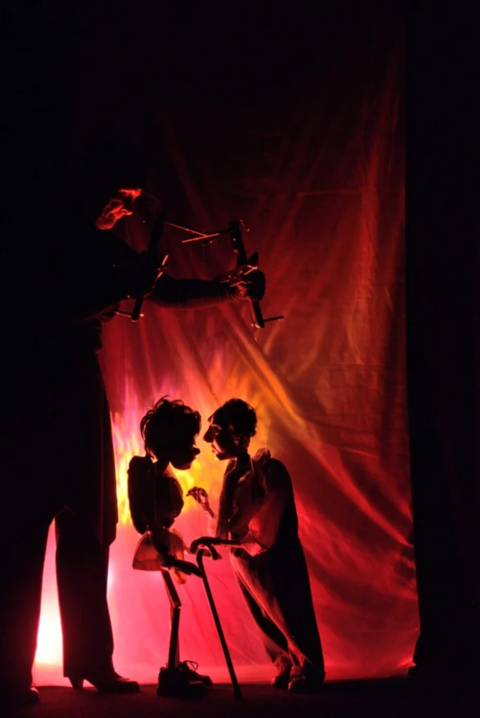 « Marionnettiste en noir manipulant deux marionnettes à fils de profil qui semblent discuter devant un rideau rouge illuminé dans une scène de théâtre d’ombres.