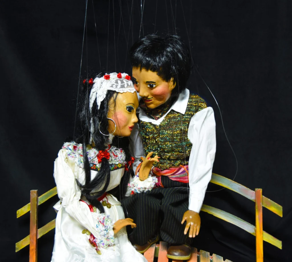 Scène tendre entre deux marionnettes à fils : la mariée en tenue blanche repose sa tête sur l’épaule du jeune homme, assis côte à côte sur un pont.