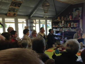 Groupe d'adultes attentifs en train de regarder Liliane Plouzeau donnant des explications sur la fabrication d'un moule en plâtre.Dans l'atelier de la Compagnie Lily : Une table, des étagères avec pots de peinture, de grandes fenêtres.