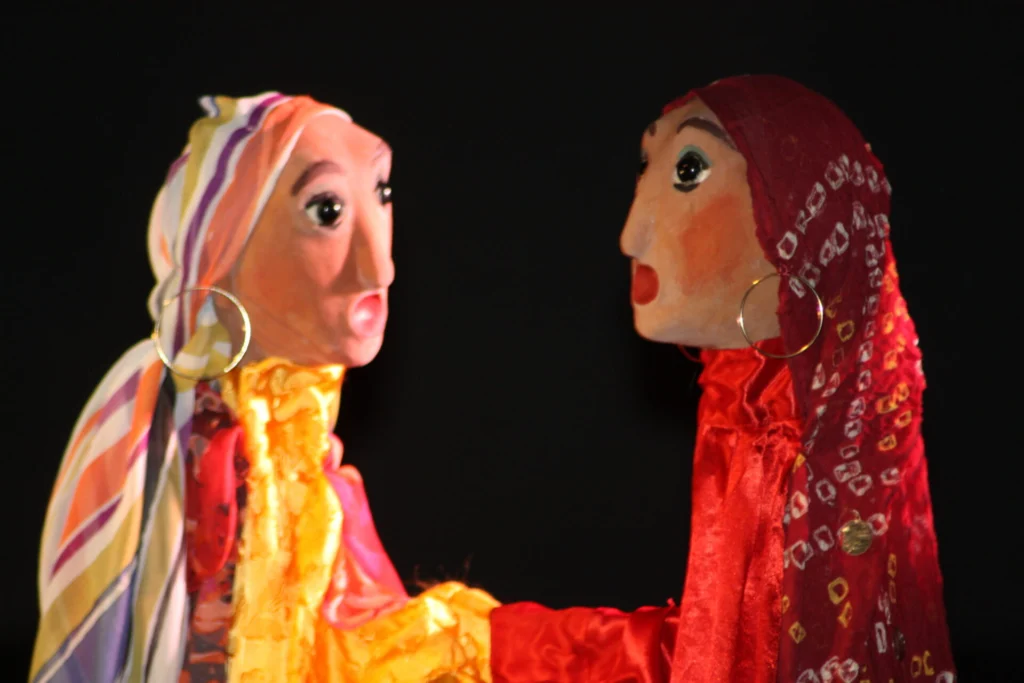 Portait serré de deux visages de marionnettes tziganes. L'une est vêtue d'un chemisier avec un foulard rouge, sur la tête. L'autre porte un chemisier jaune vif, un foulard sur la tête jaune orangé. Elles portent des anneaux aux d'oreilles.