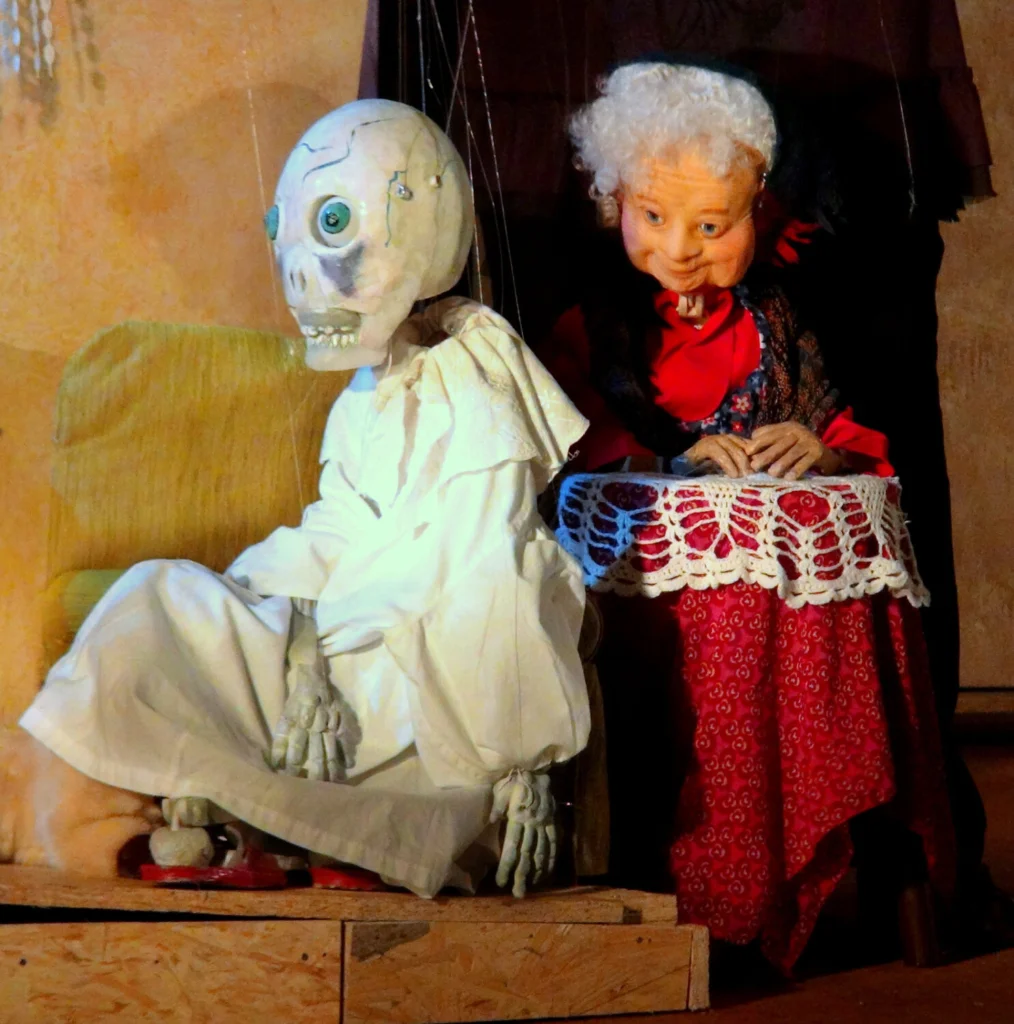 La Mort Malade, en chemise de nuit blanche, assise de profil, écoute Madeleine qui lui parle, les mains sur une petite table posée derrière la Mort Malade.