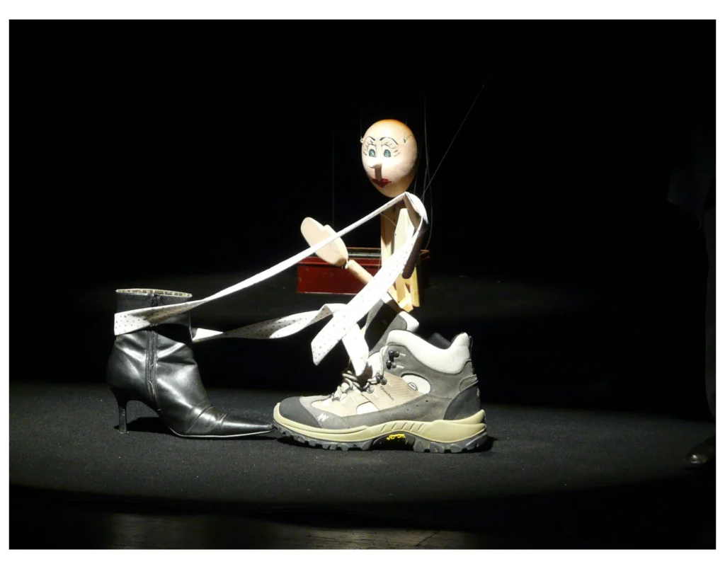 Tim, style Pinocchio amène un ruban blanc de la bottine noire vers la basket grise. Le tout sur fond noir. Visu d'une boite rouge au fond. Rond de lumière sur la scène.