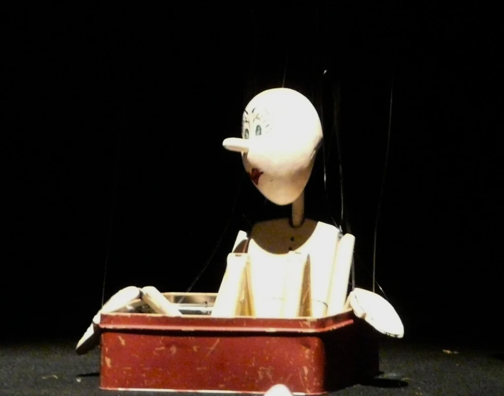 Tim, la marionnette avec son nez pointu, est assis dans une boite rouge, les mains posées sur le rebord.