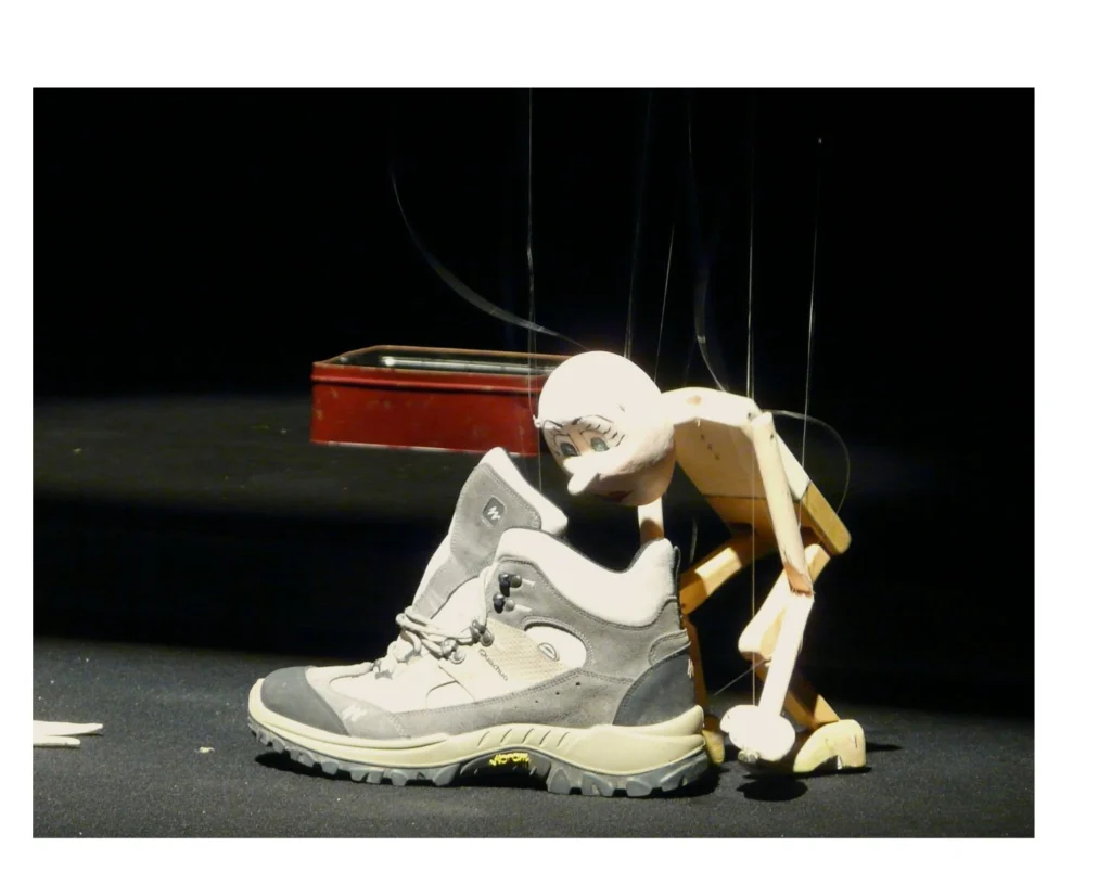 La marionnette intemporelle, style Pinocchio, sans vêtements, en bois peint beige clair pour le buste, doré en dessous, regarde l'intérieur d'une grosse chaussure de rando grise. Derrière lui, une boite rouge.