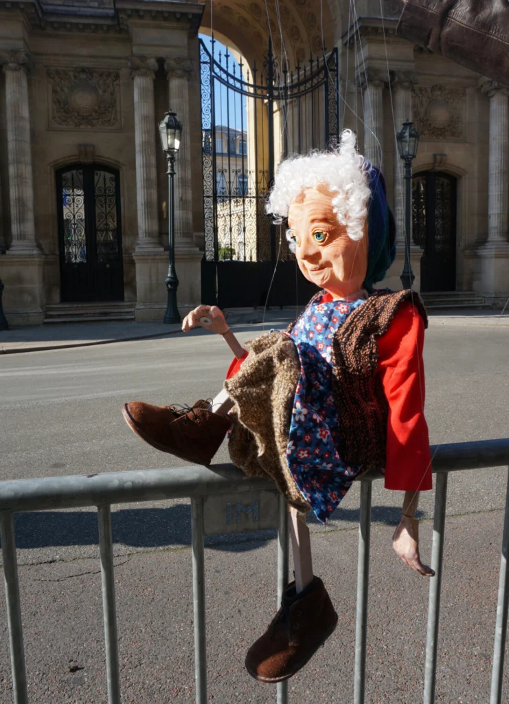 La vieille paysanne, Madeleine, marionnette à fils pose devant l'Elysée à Paris, sur une barrière grise. Elle est vêtue de sa vieille jupe marron, de son chemisier rouge et de son gilet sans manches.